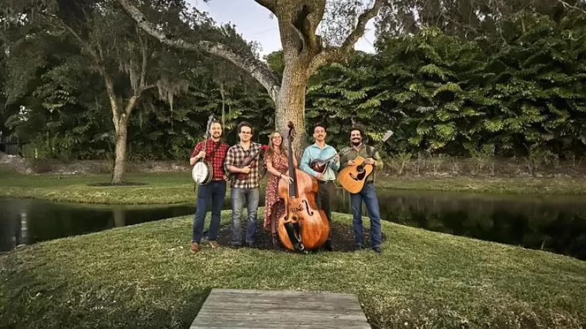 group of five people, four men and one female in the middle, holding banjos, violins, guitars and a stand up bass standing on grass infront of a lake