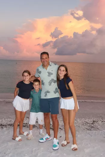The parents and three children laughing and hugging on a sandy beach at sunset, with gentle waves lapping the shore and a vibrant sky overhead.
